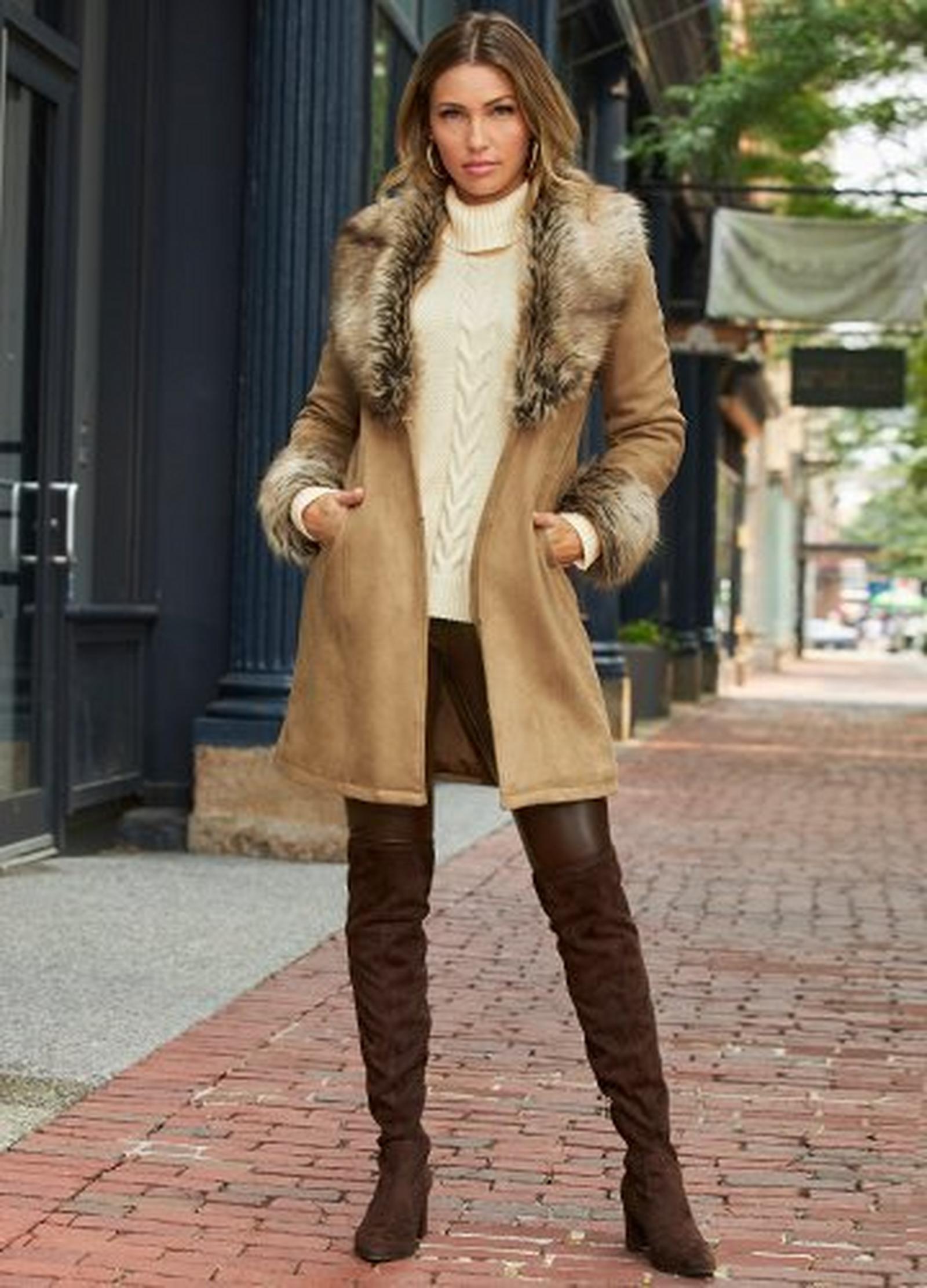 model wearing a tan suede faux-fur coat, white cable knit turtleneck sweater, brown faux-leather leggings, and brown over-the-knee suede boots.