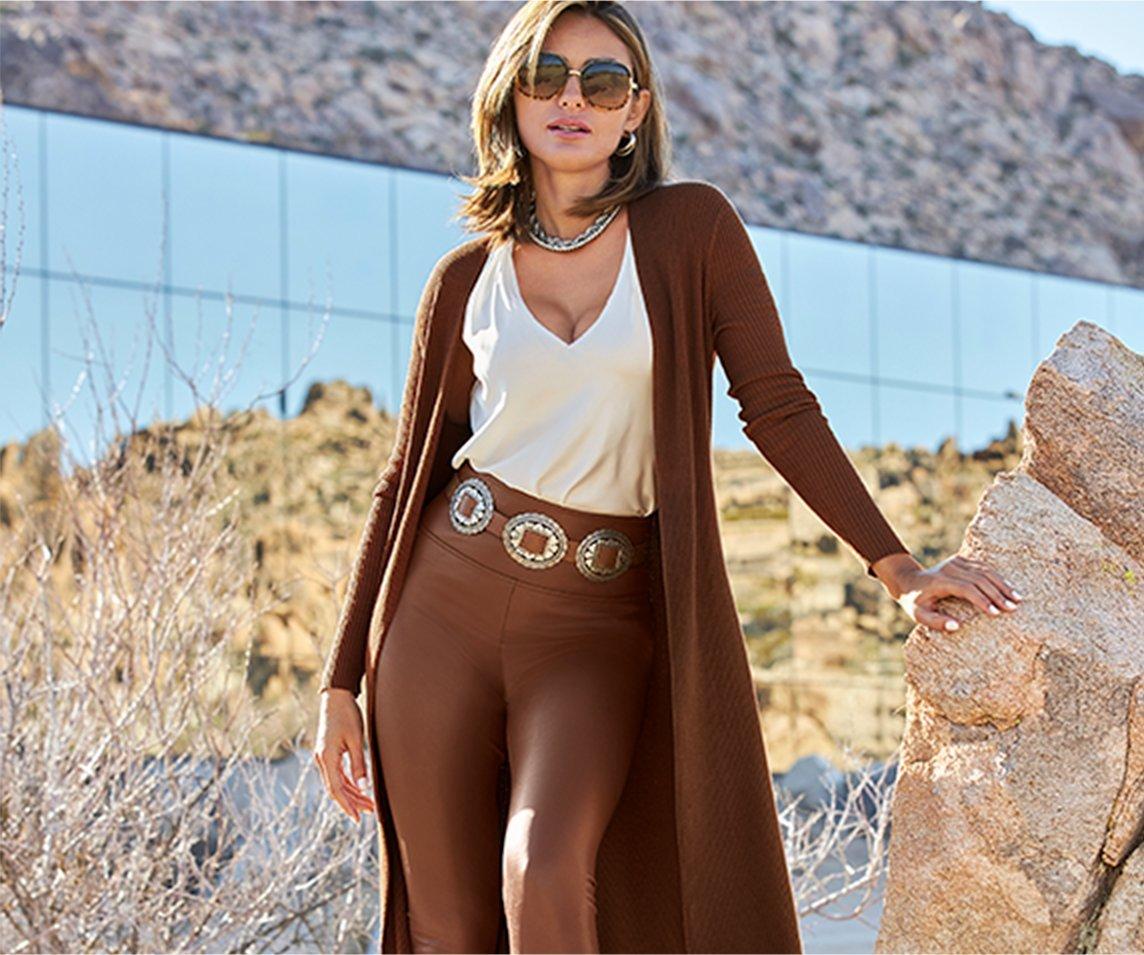 model wearing a brown long duster, off-white v-neck blouse, brown faux leather leggings, brown belt, and sunglasses.